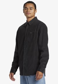 Camisa de pana negra con diseño de botones, mangas largas, bolsillo en el pecho y textura sutil, combinada con pantalones de mezclilla azul claro.