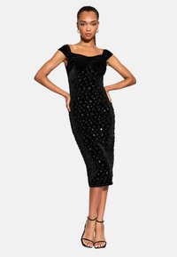 Žena oblečená v tesne priliehavých midi šatách čiernej farby s flitrami a rukávmi do polovičky, pózujúca s rukami na páse a obutá v čiernych sandáloch na členkovom opätku.