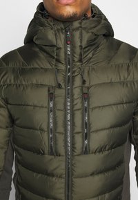 Veste olive verte rembourrée avec capuche, dotée de poches zippées, d'un tissu texturé et de panneaux latéraux gris contrastants.