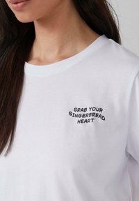 T-shirt en coton blanc avec un texte brodé en noir "SAISIS TON COEUR EN PAIN D'ÉPICE" sur la zone gauche de la poitrine. Design à manches courtes et col rond.