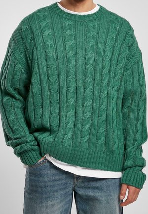 Grüner Kabelstrickpullover mit geripptem Halsausschnitt und Bündchen, im lockeren Schnitt und mit strukturiertem Muster. Über einem weißen Hemd getragen.