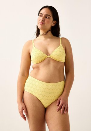 Kvinde med langt mørkt hår iført en gul, struktureret bikini-top og højtaljede underdele, stående mod en ensartet lys baggrund.