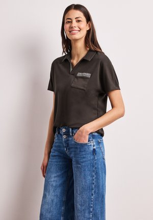 Smilende kvinde med langt brunt hår iført sort kortærmet skjorte med krave og lomme samt blå højtaljede jeans, hånd i lommen.