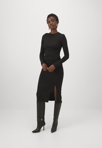 Melnas garās piedurknes kleitas ar pieguļošu augšu un sānu šķēlumu, kombinēta ar melniem augstiem apkaklīšu zābakiem. Gluda auduma, minimālistisks dizains.