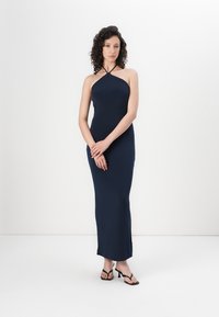 Mørkeblå halterneck maxikjole, figurnær design, glatt stoff, ankellengde, kombinert med svarte sandaler med stropper. Enkel og elegant.