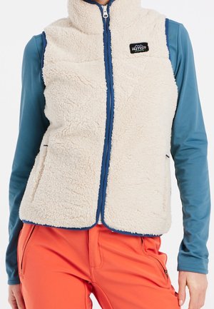 Person wearing beige fleece vest with navy zipper and logo, blue long sleeve shirt, and bright orange pants.