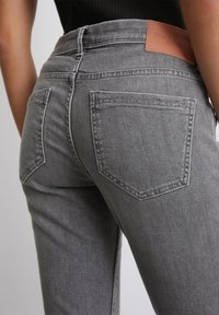 Un pantalon en denim gris présente un design ajusté avec une taille haute, cinq poches et une étiquette en cuir sur la ceinture arrière.
