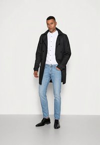 Czarny płaszcz trencz na białej koszuli, w połączeniu z jasnoniebieskimi jeansami i czarnymi butami. Płaszcz ma dwurzędowy fason i guziki.