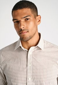 Camicia chiara con bottoni a maniche lunghe, caratterizzata da un motivo geometrico marrone. Presenta un colletto a punta e un tessuto texturizzato. Design semplice e su misura.