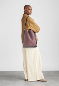 Lila táska fekete díszítéssel, sima anyagból készült, minimalista dizájnnal és egyetlen vállpánttal rendelkezik. A modellen semleges színű nadrág és felső van.