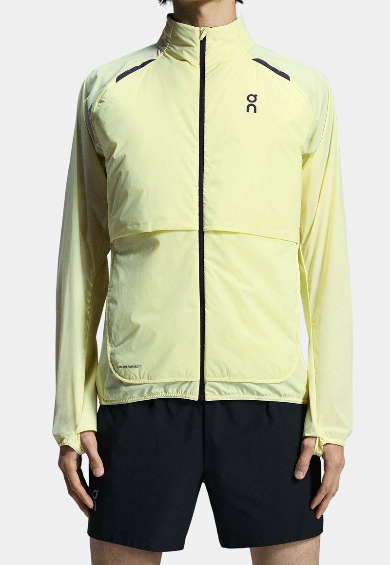 Leichte gelbe Laufjacke mit Reißverschluss vorne, elastischen Bündchen und schwarzen Akzenten. Verfügt über einen breiteren Oberkörper und ein Logo auf der Brust.