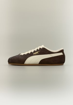 Brauner Puma-Sneaker aus Wildleder mit cremefarbigem Seitenstreifen, cremefarbenen Schnürsenkeln und goldenem Puma Tackle-Logo, seitlich auf neutralem Hintergrund dargestellt.