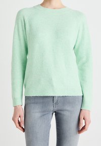 Kvinna klädd i en ljusgrön stickad tröja och ljusgrå tajta jeans, stående mot en enkel vit bakgrund.