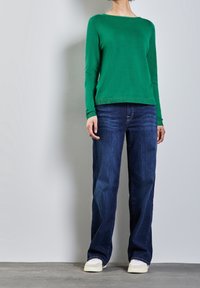 Pull en tricot vert à manches longues, col rond, associé à un jean droit bleu foncé et des baskets de couleur claire sur un sol gris.