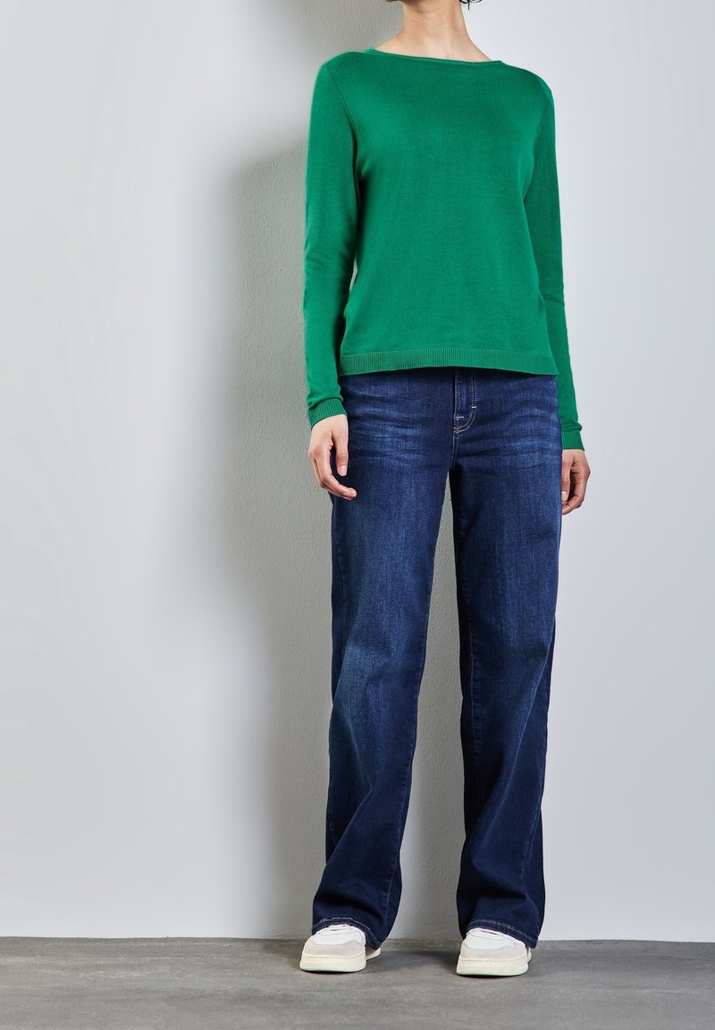 Pull en tricot vert à manches longues, col rond, associé à un jean droit bleu foncé et des baskets de couleur claire sur un sol gris.