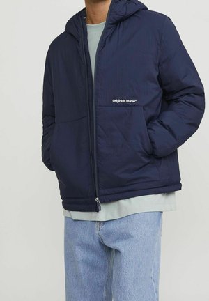 Osoba nosząca granatową puchową kurtkę z kapturem z napisem "Originals Studio" na klatce piersiowej, jasno-szarą koszulę i jasno-niebieskie dżinsy.