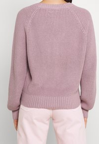 Kvinna iklädd en långärmad, lila stickad tröja och ljusrosa byxor, sedd bakifrån mot en enfärgad bakgrund.