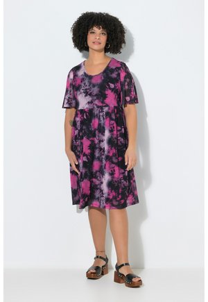 Frau mit lockigem Haar, die ein knielanges, kurzärmeliges, pink-schwarzes Batik-Kleid und schwarze Plateausandalen trägt, steht vor einer weißen Wand.