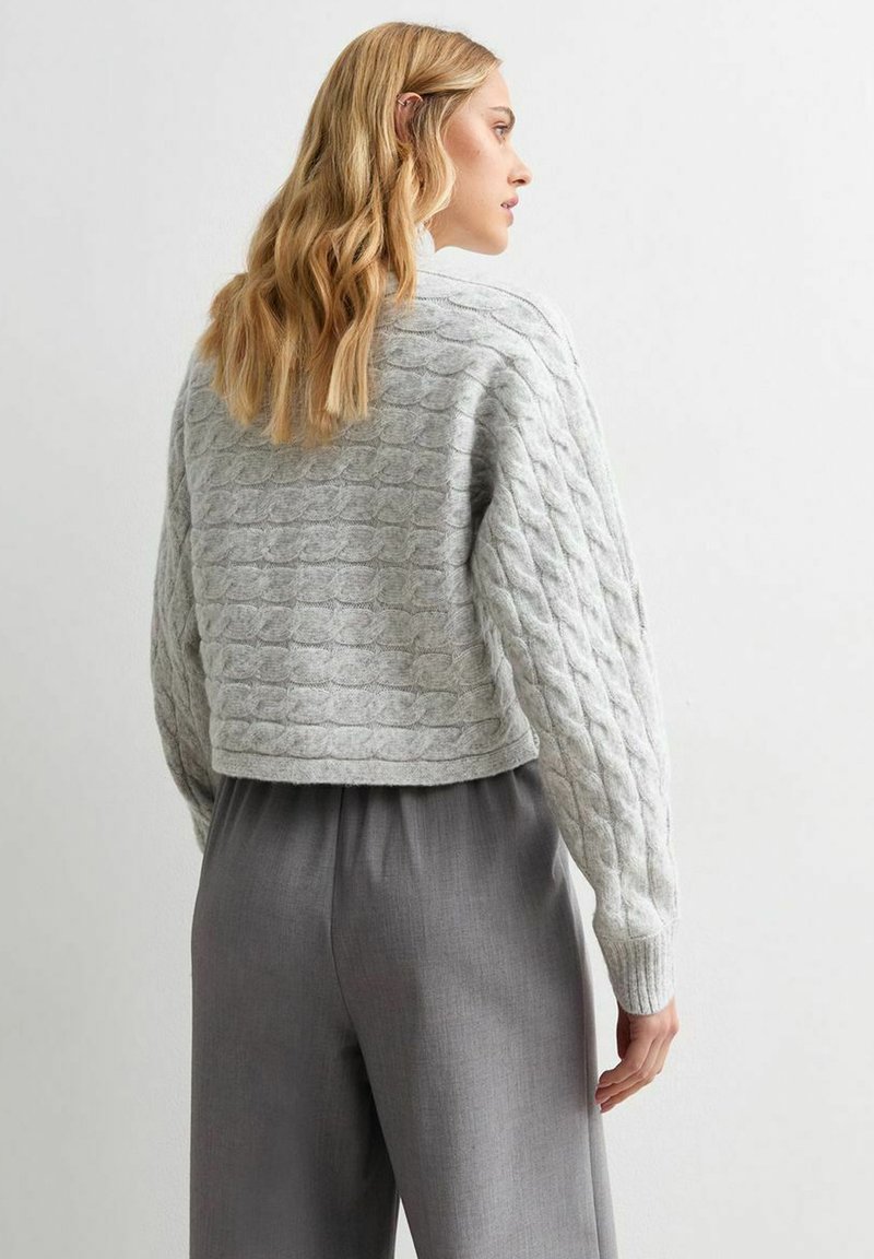 Maglione grigio chiaro cropped con un motivo a trecce testurizzate; presenta maniche lunghe e polsini a coste, abbinato a pantaloni grigi a gamba larga.
