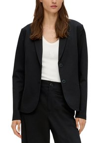 Vrouw in een zwarte blazer en bijpassende broek over een witte tanktop met een subtiele logo op de borst.