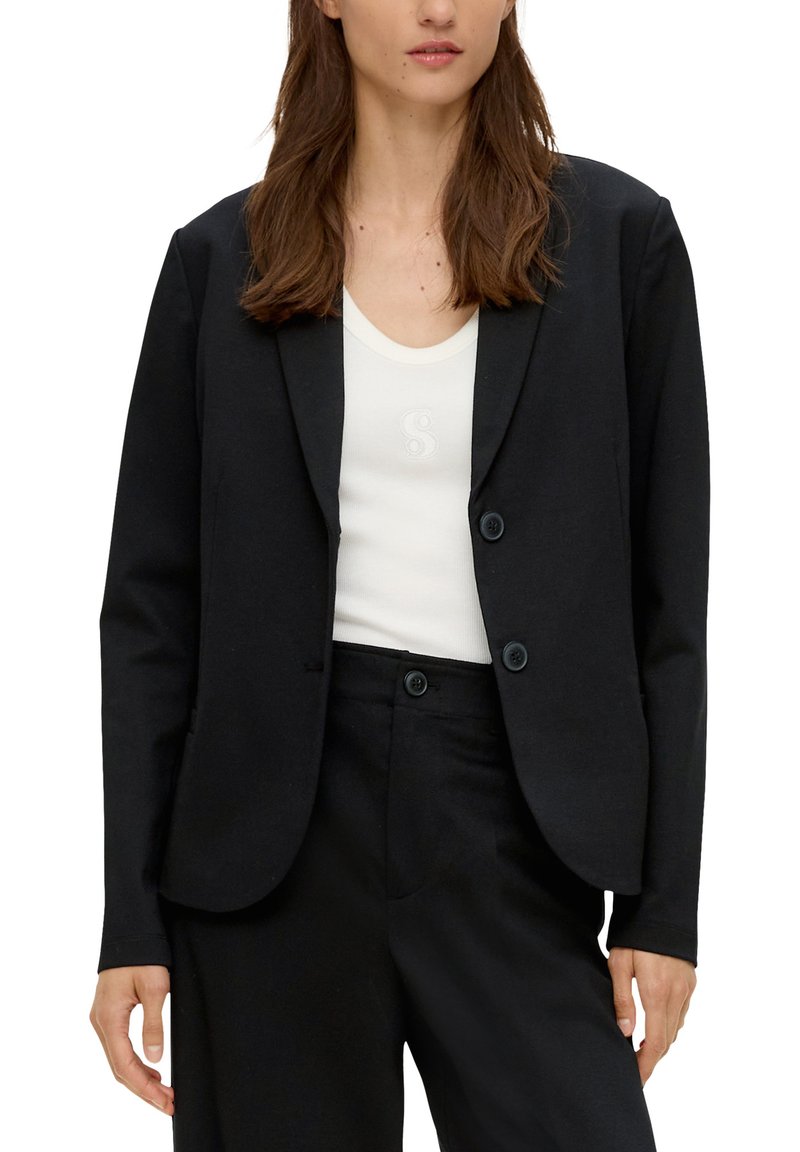 Femme portant un blazer noir et un pantalon assorti sur un débardeur blanc avec un logo discret sur la poitrine.