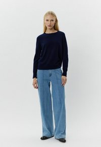 Mornarska pulover s okruglim izrezom, uparen s svijetloplavim širokim trapericama i crnim cipelama s srebrnim akcentima. Postavljen na jednostavnoj pozadini.