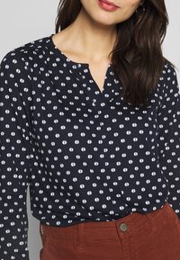 Marinebluse mit weißem Halbkreis-Muster, V-Ausschnitt und langen Ärmeln; getragen mit rostfarbenen Cordhosen. Glatte Textur.