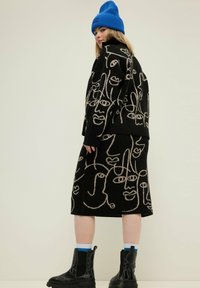 Schwarzes Strickoutfit mit abstrakten Gesichts-Mustern in Beige, kombiniert mit einer leuchtend blauen Mütze und schwarzen Stiefeletten mit dicken Sohlen.