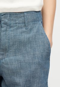 Κοντινό πλάνο μπλε τζιν παντελονιού με ορατή θηλιά ζώνης και πλαϊνή τσέπη, φορεμένο με λευκό ριμπ τοπ που είναι κρυμμένο μέσα στο παντελόνι.