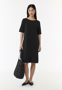 Robe noire mi-longue à manches courtes, col rond, coupe droite. Le modèle porte un sac matelassé noir et des ballerines noires à bout pointu.