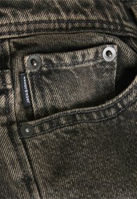 Μαύρα τζιν παντελόνια σε κοντινή λήψη, με υφή ύφασματος, ορατές ραφές, δύο μεταλλικά κουμπιά και ετικέτα μάρκας στην μπροστινή τσέπη.