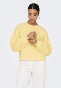 Vrouw met donker haar, die grote hoepeloorbellen draagt, een gele trui en een witte broek, kijkt naar beneden terwijl ze haar ringen aanpast tegen een effen achtergrond.