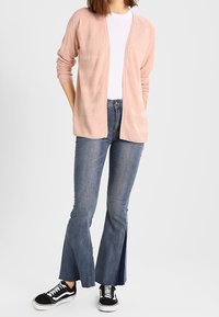 Cardigan rosa chiaro con un motivo testurizzato, indossato su una camicia bianca. Abbinato a jeans svasati scuri e sneakers nere con dettagli bianchi.