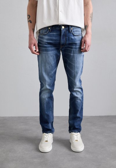 Jeans in denim blu sbiadito con taglio dritto, cinque tasche e dettagli in rame, abbinati a sneaker bianche. Sfondo semplice.