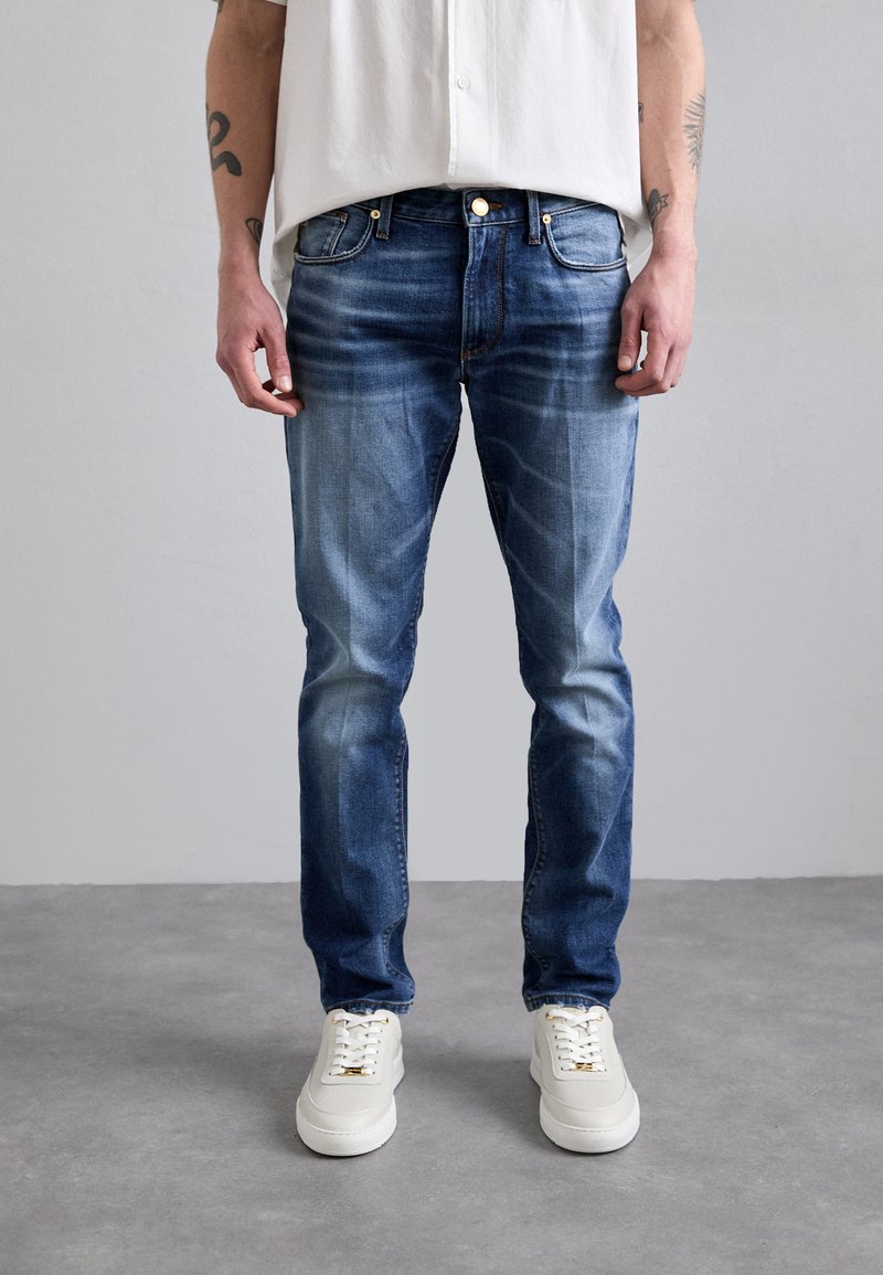 Jeans din denim albastru estompat, cu o croială dreaptă, cinci buzunare și accesorii din cupru, asociate cu pantofi sport albi. Fundal simplu.