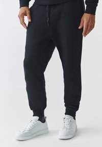 Sorte joggers med sidelommer, elastiske manchetter og snor liv, parret med hvide sneakers og en glat tekstur.