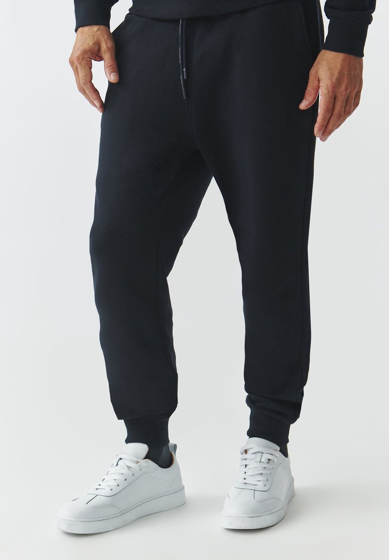 Sorte joggers med sidelommer, elastiske manchetter og snor liv, parret med hvide sneakers og en glat tekstur.