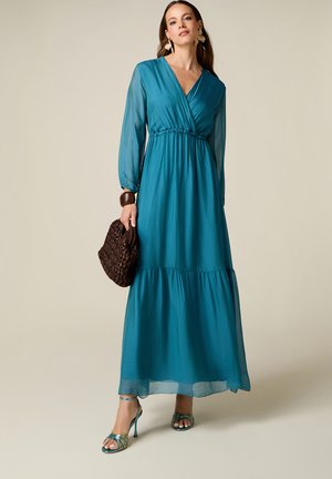 Femme en robe longue maxi transparente à manches longues couleur bleu sarcelle, tenant un sac à main tressé foncé, portant des talons à brides bleu sarcelle, de grandes boucles d'oreilles et un bracelet foncé.