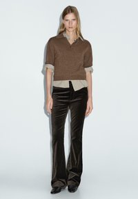 Bruine gekropte trui met een kraag over een lichtbeige shirt, gecombineerd met donkerbruine fluwelen flare-broeken en zwarte schoenen.