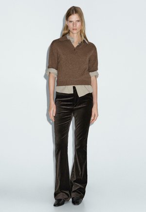 Maglione cropped marrone con colletto, sovrapposto a una camicia beige chiaro, abbinato a pantaloni a zampa in velluto marrone scuro e scarpe nere.