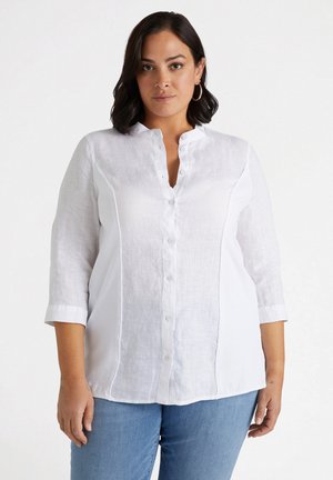 Blouse en lin blanc avec col mandarin, devant à boutons, et manches trois-quarts, présentant un tissu texturé subtil.