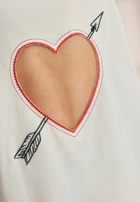 Witte kleding met een hartvormige uitsnijding omrand met rode stiksels, met een zwart pijltje dat het hart kruist.