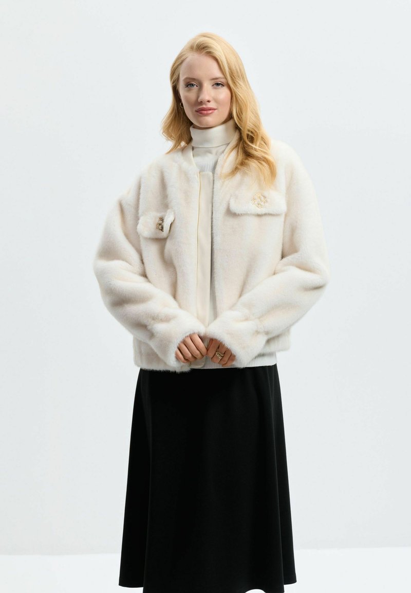 Chaqueta bomber blanca de felpa con una textura suave, dos bolsillos frontales y un detalle dorado. Lleva puesta encima de un suave suéter de cuello alto y una falda negra.