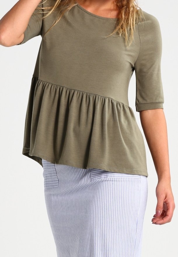 Femme portant un top péplum vert olive à manches mi-longues associé à une jupe bleu clair à fines rayures blanches, posant avec un bras levé.