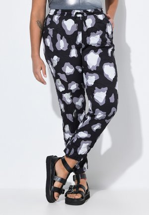 Pantalones negros con un patrón que presenta formas abstractas en gris y blanco, con una cintura ajustable y un corte holgado. Se llevan con sandalias negras gruesas.