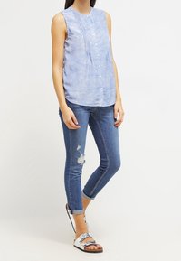 Blusa senza maniche azzurro chiaro con chiusura a bottoni e pieghe verticali, abbinata a jeans blu attillati con accenti degradè e orli arrotolati. Sandali argentati.