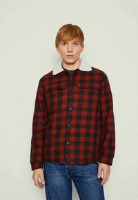 Jovem vestido com uma jaqueta xadrez vermelha e preta com colarinho de lã branca e jeans azuis, encostado a um fundo claro.