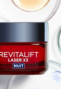 Bocal en verre rouge avec un couvercle noir, étiqueté "REVITALIFT LASER X3 NUIT." Entouré de contenants transparents circulaires contenant divers liquides colorés.