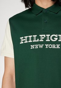 Nahaufnahme eines grünen Poloshirts mit weißen kurzen Ärmeln und dem aufgestickten Schriftzug "HILFIGER NEW YORK" auf der Brust.