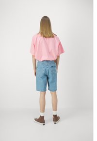 Rosa oversized skjorta, blå denimshorts och bruna högre sneakers med vita strumpor. Enhetlig bakgrund, fokus på avslappnad outfit.